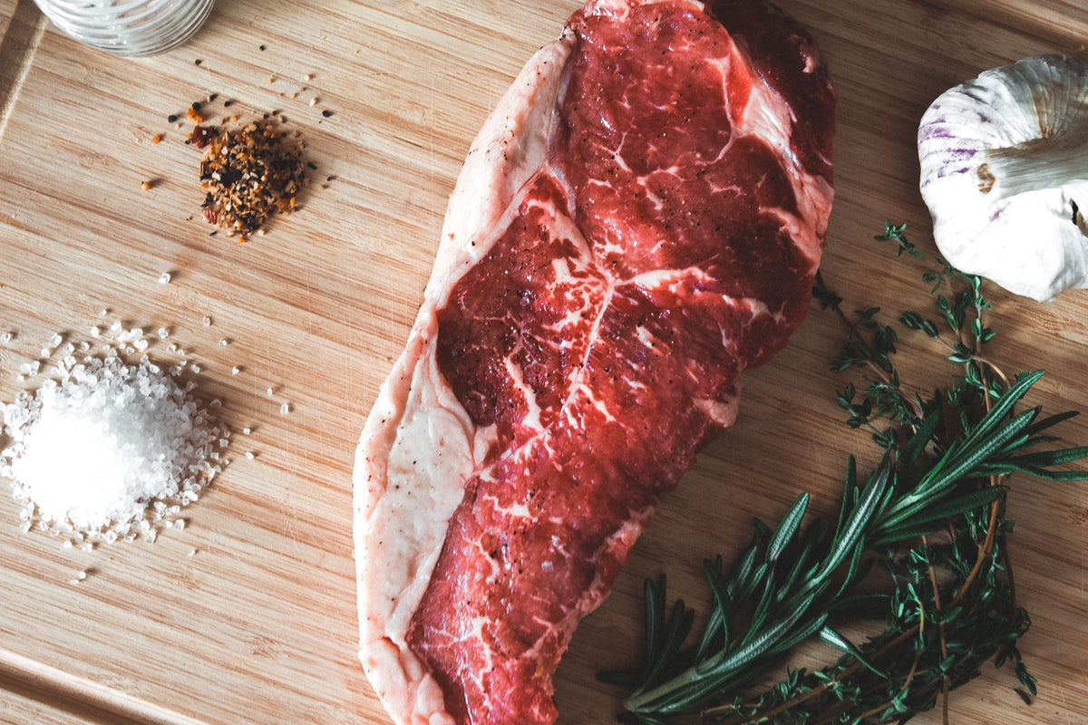 A grass-fed New York strip on a wooden board with salt, garlic and rosemary